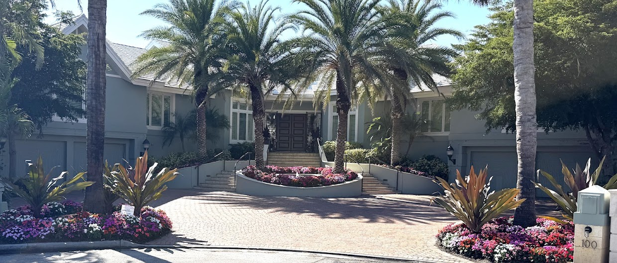 Front elevation of a Sarasota coastal estate: pale green residence framed by mature palms, with paver motor court and seasonal flower beds in bloom.